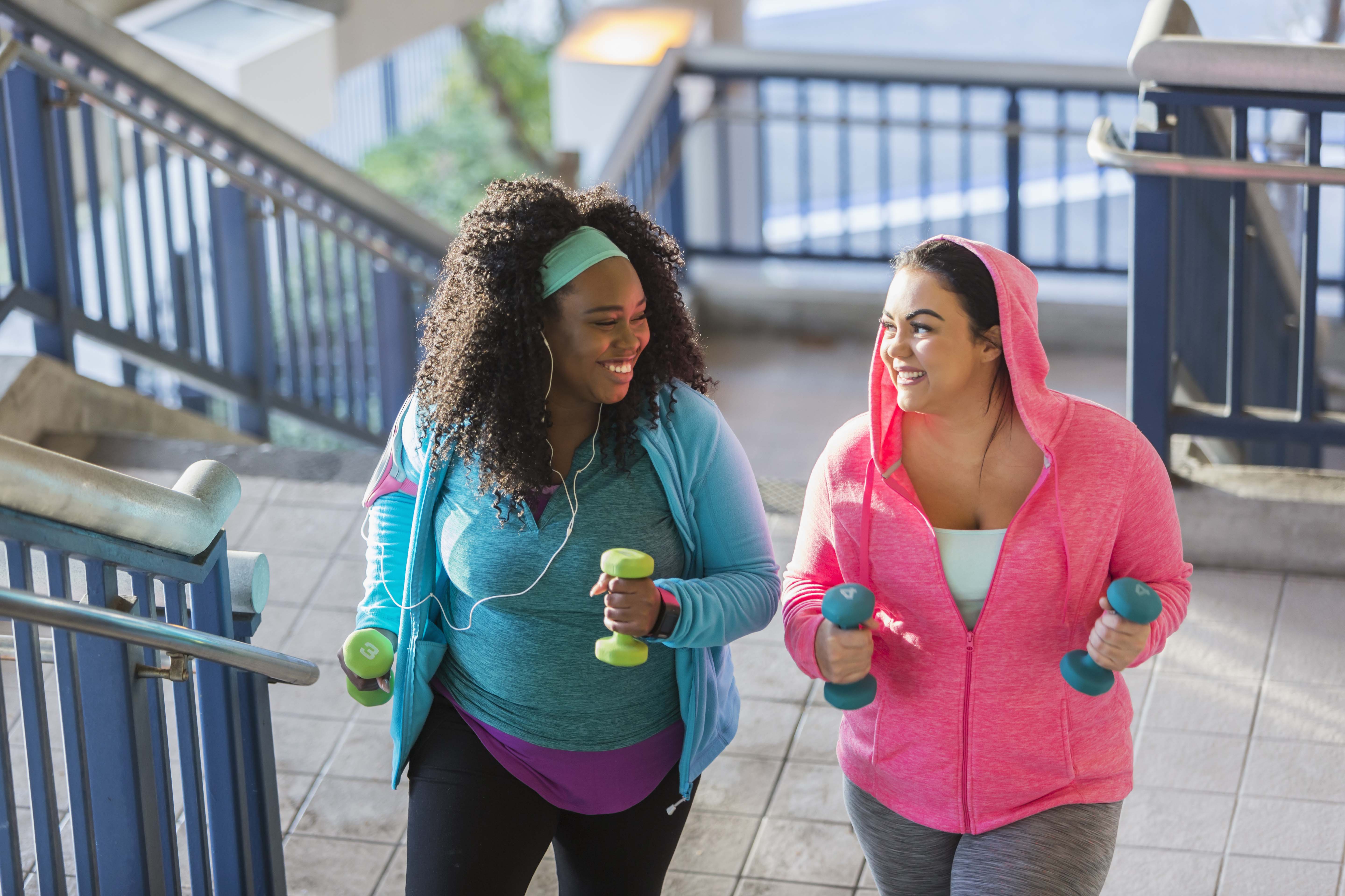 Two multi-ethnic young women exercising together. They are looking at each other, smiling, as they climb a staircase holding hand weights.